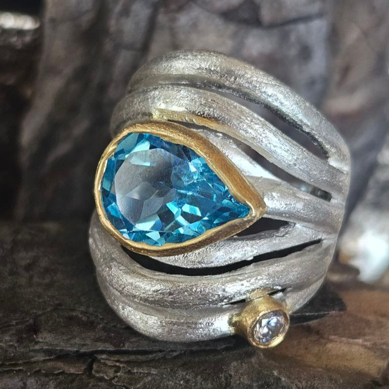 A silver ring featuring a large blue topaz stone with gilding details and small clear stones set in a textured band.